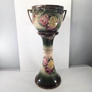 Puede incluir: Una jardinera de cerámica antigua con base de pedestal. La jardinera presenta rosas pintadas a mano en tonos rosa y amarillo sobre un fondo crema. El pedestal es verde y marrón con detalles florales. La jardinera tiene dos asas.