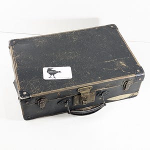 May include: A rectangular black vintage suitcase with a weathered, textured surface, metal corner reinforcements, and brass-toned latches. A white rectangular sticker featuring a black crow silhouette and the text 1000crows is attached.