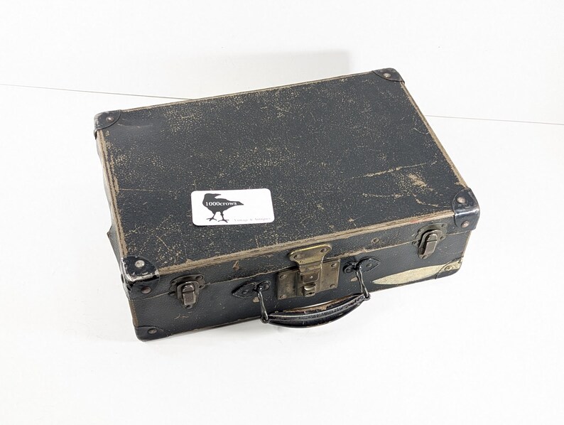 May include: A rectangular black vintage suitcase with a weathered, textured surface, metal corner reinforcements, and brass-toned latches. A white rectangular sticker featuring a black crow silhouette and the text 1000crows is attached.