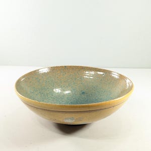 May include: A round ceramic bowl featuring a smooth, mustard-yellow exterior with a subtle horizontal line detail. The interior is finished with a speckled, mottled glaze in shades of teal and light blue, creating a textured appearance.