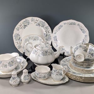 Könnte beinhalten: Ein komplettes Vintage-Teeservice mit weißem und hellblauem Blattmuster. Das Set umfasst Teller, Tassen, eine Teekanne, Sahnekännchen, Zuckerdose sowie Salz- und Pfefferstreuer. Die Ränder der Teller und Tassen sind goldfarben.