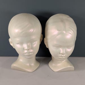 May include: Two off-white, child-sized mannequin heads. The heads have detailed facial features, including closed eyes and a slightly downturned mouth. The hair is styled with a side part. The heads are displayed on a light gray surface.