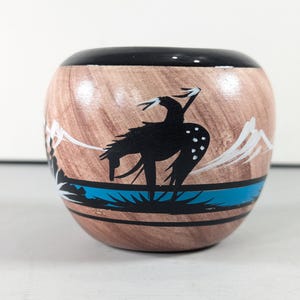 May include: A round, wooden decorative bowl with a black rim and a brown wood grain pattern. The bowl features a black silhouette of a bird with white accents, a blue stripe, and white mountain designs.