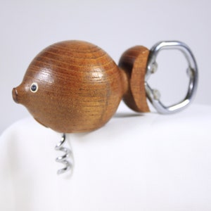 May include: A wooden fish-shaped corkscrew with a silver metal spiral and bottle opener. The fish has a round body, a small snout, and a single eye. The wood has a natural grain and a warm brown color.