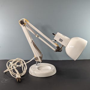 May include: A white desk lamp with an adjustable arm and a rounded lampshade. The lamp has a circular base and a white power cord. The lamp's arm is made of metal and has a series of joints that allow it to be positioned in various ways.