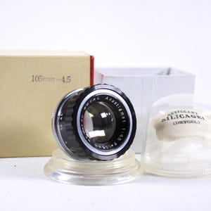 Op de afbeelding: Een vintage cameralens met een zwart en zilverkleurig lichaam, rustend op een heldere plastic standaard. De lens wordt vergezeld door een kartonnen doos met "105mm = 4.5" erop gedrukt, en een doorzichtige plastic container met een "SILICAGEL"-zakje.