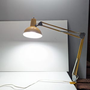 May include: A yellow and silver articulated desk lamp with a bright light. The lamp has a rounded shade and a flexible arm, and is connected to a power cable. The lamp is on a white surface.