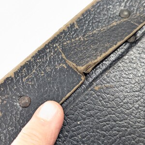 May include: A black textured surface with a worn beige trim edge, featuring visible metal rivets along the border. The material shows signs of age with frayed edges and surface wear, indicating a vintage construction with a pebbled leather-like finish.