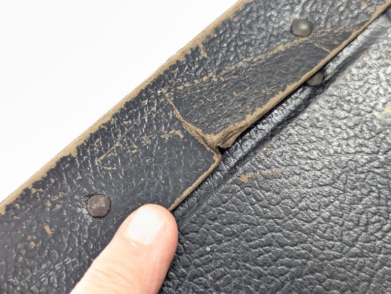 May include: A black textured surface with a worn beige trim edge, featuring visible metal rivets along the border. The material shows signs of age with frayed edges and surface wear, indicating a vintage construction with a pebbled leather-like finish.