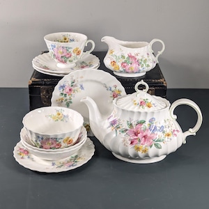 Puede incluir: Un juego de té de porcelana blanca con motivos florales. El juego incluye una tetera, una jarra de crema, una taza de té, un platillo y un cuenco con un platillo a juego. El diseño floral presenta flores rosas, amarillas y azules con hojas verdes.