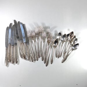 May include: A collection of vintage silver cutlery, comprising knives, forks, and spoons, displayed on a white surface. The silverware features a silver finish with ornate handle detailing. The arrangement suggests a formal dining setting.