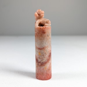 May include: A cylindrical, carved stone seal with a small animal figure on top. The seal is primarily a mix of pink, red, and cream colors, with a marbled appearance. The animal figure is a light pink color. The seal is standing upright on a white surface.