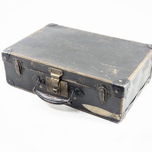 May include: A rectangular black suitcase with a worn, textured surface. It features brass-toned metal latches, a central locking mechanism, and reinforced metal corner protectors. A black handle is attached to the front with metal hardware.