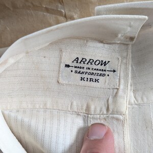 May include: Close-up of a cream-colored shirt collar with a label that reads "ARROW MADE IN CANADA SANFORIZED KIRK". The shirt has vertical stripes and a buttoned placket.