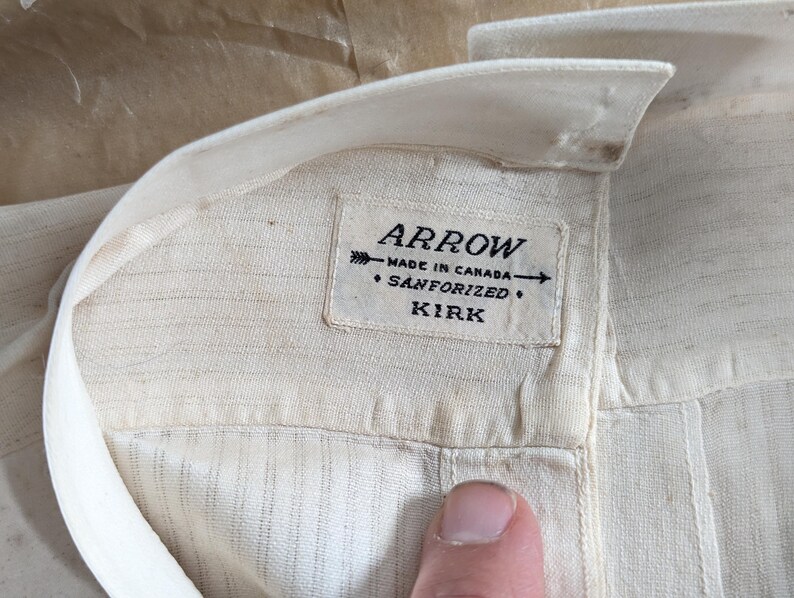May include: Close-up of a cream-colored shirt collar with a label that reads "ARROW MADE IN CANADA SANFORIZED KIRK". The shirt has vertical stripes and a buttoned placket.