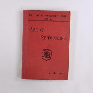 Puede incluir: Un libro rojo vintage titulado "Art of Re-touching" de "The Amateur Photographer Library". La portada presenta texto negro y un emblema decorativo. El nombre del autor, J. Hubert, está en la parte inferior.