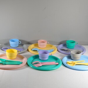 May include: A set of six pastel-colored plastic plates, cups, and utensils. The plates are round and have a slightly raised edge. The cups are cylindrical and have a handle. The utensils are forks and spoons. The colors are light blue, light green, light pink, yellow, and gray.