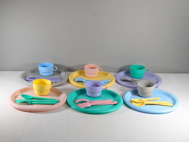 May include: A set of six pastel-colored plastic plates, cups, and utensils. The plates are round and have a slightly raised edge. The cups are cylindrical and have a handle. The utensils are forks and spoons. The colors are light blue, light green, light pink, yellow, and gray.