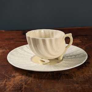 Puede incluir: Juego de taza y platillo de té color crema. La taza tiene un diseño acanalado y un asa curva. El platillo tiene un patrón texturizado a juego. El juego es de cerámica.