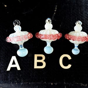 May include: Three glass ornaments with a white top, a pink and red frilly midsection, and a light blue base. Each ornament has a loop at the top for hanging. Wooden letters A, B, and C are below the ornaments.