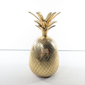 May include: A gold-colored pineapple-shaped decorative object. The pineapple has a textured, diamond-patterned body and a spiky crown. The object is likely made of metal and is designed for decorative purposes.