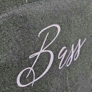 May include: Close-up of a dark green fabric with the cursive word "Bass" in white. The fabric has a textured appearance, and the lettering is elegantly styled.