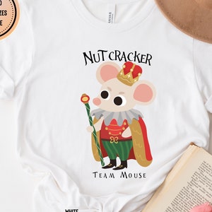 May include: White t-shirt featuring a cartoon mouse dressed as a Nutcracker character, wearing a crown and holding a staff. The text "NUTCRACKER TEAM MOUSE" is above and below the illustration. The words "ASSORTED COLORS + SIZES AVAILABLE" are in a circle.