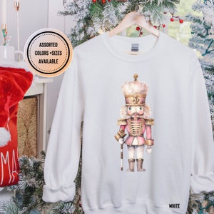 May include: White sweatshirt featuring a watercolor illustration of a pink and gold nutcracker soldier. The design includes a detailed depiction of the nutcracker's uniform and facial features. The text "WHITE" is at the bottom right. "ASSORTED COLORS + SIZES AVAILABLE" is in a circle.
