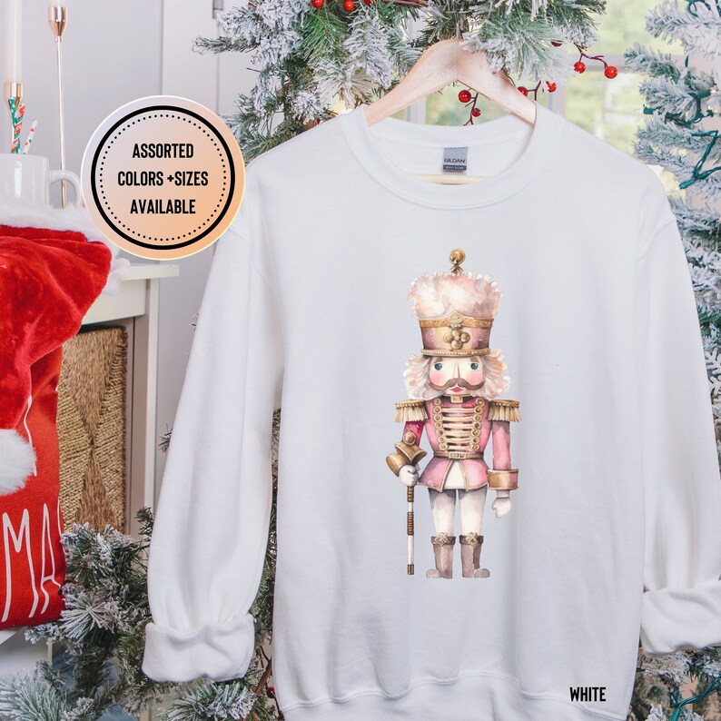 May include: White sweatshirt featuring a watercolor illustration of a pink and gold nutcracker soldier. The design includes a detailed depiction of the nutcracker's uniform and facial features. The text "WHITE" is at the bottom right. "ASSORTED COLORS + SIZES AVAILABLE" is in a circle.