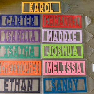 May include: A collection of colorful name tags in various colors, including yellow, blue, red, purple, green, orange, pink, and gray. Each tag features a name cut out in a bold font. The names include KAROL, CARTER, EMMANUEL, ISABELLA, MADDIE, ISATHA, JOSHUA, CHRISTOPHER, MELISSA, ETHAN, and SANDY.