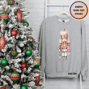 May include: Sport grey sweatshirt featuring a watercolor illustration of a pink and gold nutcracker soldier. The text "ASSORTED COLORS + SIZES AVAILABLE" is in a circle at the top right. The words "SPORT GREY" are printed at the bottom. A Christmas tree with ornaments is in the background.