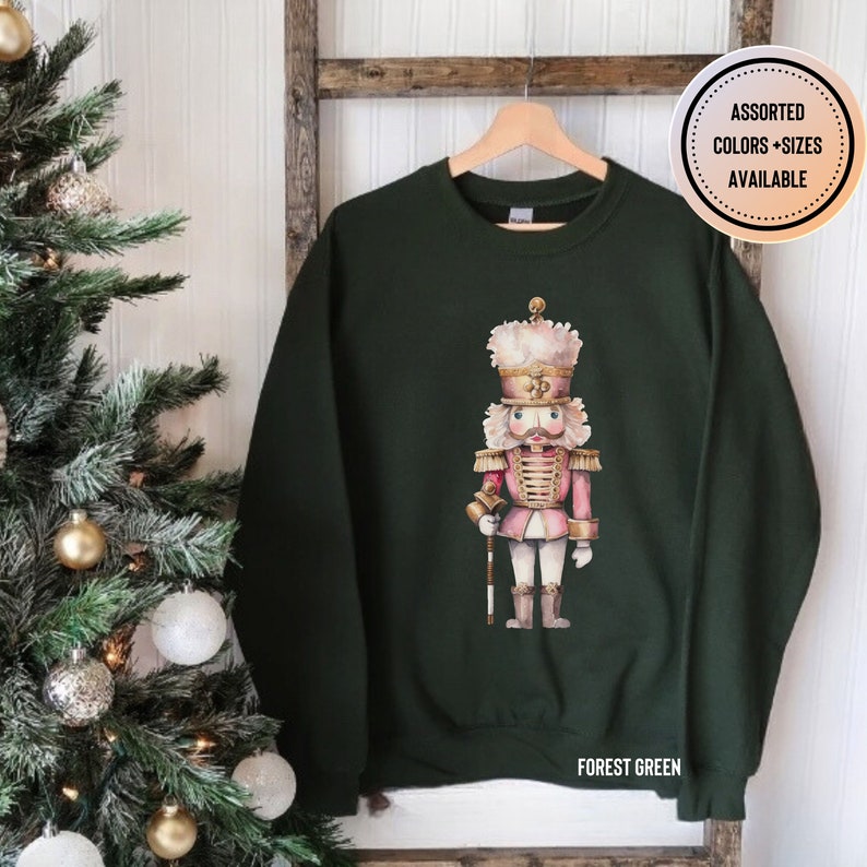 May include: Forest green sweatshirt featuring a watercolor illustration of a pink and gold nutcracker. The nutcracker is wearing a tall hat and holding a staff. The text "ASSORTED COLORS + SIZES AVAILABLE" is in a circle on the upper right.