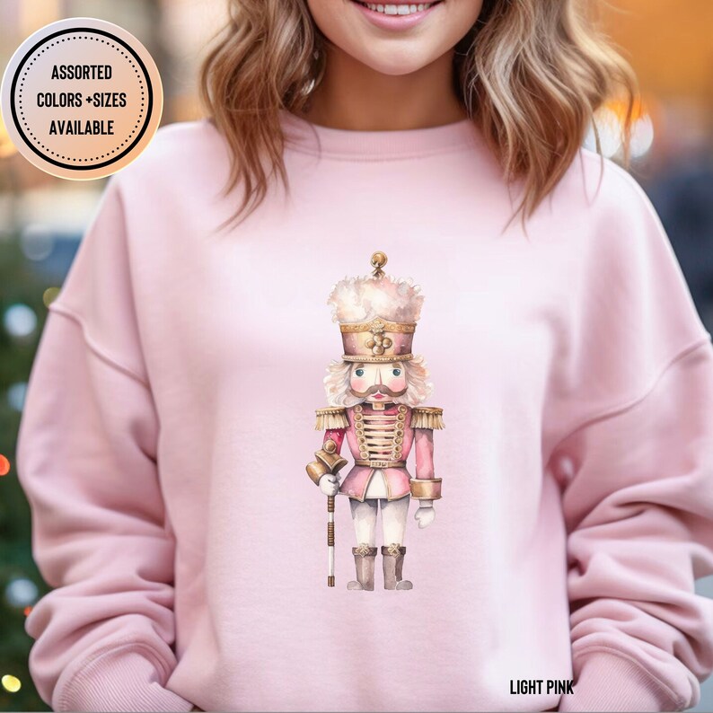 May include: Light pink sweatshirt featuring a watercolor illustration of a Nutcracker soldier. The Nutcracker is in a pink and gold uniform, holding a staff. The text "ASSORTED COLORS + SIZES AVAILABLE" is in a circle on the upper left. The text "LIGHT PINK" is at the bottom.