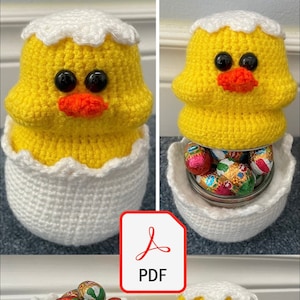 Könnte beinhalten: Ein gehäkeltes Küken mit einer weißen Eierschalenbasis. Das Küken ist gelb mit schwarzen Augen und einem orangefarbenen Schnabel. Die Eierschale ist weiß mit einem gewellten Rand. Das Küken hält ein Glas mit bunten Schokoladeneiern.