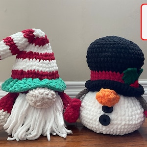 以下が含まれることがあります： 編みぐるみで作られた、ノームと雪だるまのクリスマスデコレーションセット。ノームは赤と白のストライプの帽子に緑色のつば、白いひげ、赤いミトンを付けています。雪だるまは黒いシルクハットに赤いリボンと緑のヒイラギ、オレンジ色のニンジンの鼻、赤いミトンを付けています。