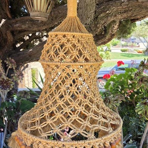 May include: A tan macrame hanging light fixture featuring a bell-shaped design with intricate knotted patterns. The lower edge is adorned with a row of smooth, round wooden beads and a thick, textured fringe made of natural fiber cord.