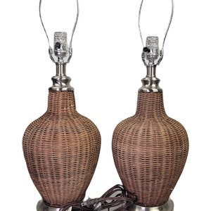 May include: A pair of wicker table lamps with a brown woven shade and a silver base. The lamps have a cord and plug.