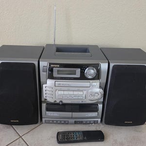 May include: A silver and black Aiwa stereo system featuring a central unit with a CD player, dual cassette decks, a digital display, and a vertical antenna. Two detachable speakers with black grilles flank the unit, accompanied by a black remote control.