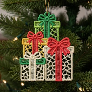 Op de afbeelding: Geborduurde kerstboomversiering met drie gestapelde cadeau dozen in groen, geel en wit. Elke doos heeft een contrasterende strik en lint. De versiering hangt aan een gouden draad aan een kerstboom.