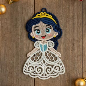 May include: Embroidered princess ornament with a blue and gold tiara, dark blue hair, and a white dress with a decorative pattern. The ornament is surrounded by gold baubles and festive lights, perfect for holiday decor.