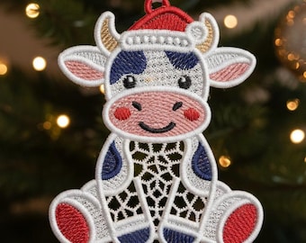 May include: Embroidered Christmas ornament shaped like a cow wearing a Santa hat. The ornament features red, white, and blue accents, with a detailed openwork design on the body. It hangs from a white ribbon against a blurred Christmas tree background.