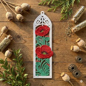 以下が含まれることがあります： 赤いポピー、緑の葉、つぼみの刺繍が施された白いフレームのポピーのデザイン。 ポピーのデザインは、乾燥した種子のさや、小枝、小さなガラス瓶のある木製の背景に配置されています。