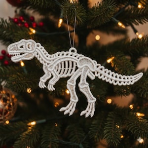 May include: A white, embroidered dinosaur skeleton Christmas ornament. The ornament is in the shape of a Tyrannosaurus Rex, with detailed stitching outlining the bones and body. It hangs from a thin, silver string against a backdrop of a Christmas tree.