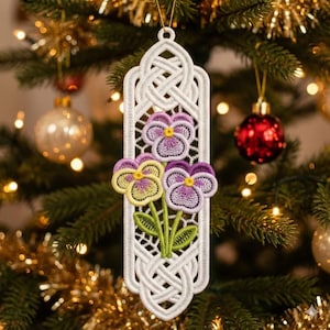 以下が含まれることがあります： 白の刺繍クリスマスオーナメント。ケルトノットのデザインで、黄色、紫、緑色のパンジーが3つ描かれています。オーナメントは長方形で、吊り下げ用のループが付いています。背景はクリスマスツリーです。