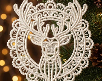 Op de afbeelding: Een witte, ingewikkeld geborduurde kerstversiering. Het ontwerp toont een rendierkop in een cirkelvormig frame, versierd met delicate, kantachtige details. De versiering hangt tegen een achtergrond van dennentakken en zachte, vage lichten.
