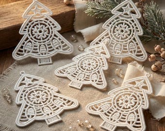 May include: Five white, intricately designed Christmas tree ornaments. Each tree features a star at the top and a detailed snowflake and tree pattern. The ornaments are placed on a neutral-colored fabric with decorative elements.