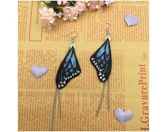 May include: A pair of blue and black butterfly earrings with silver chains. The earrings are on a brown background with pink flowers and purple hearts.