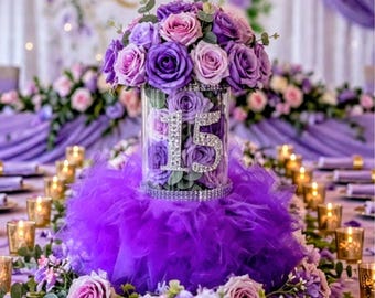 Könnte beinhalten: Eine zylindrische Glasvase gefüllt mit violetten und hellrosa Kunstrosen, die auf der Vorderseite eine mit Strasssteinen besetzte Zahl 15 zeigt. Der Fuß der Vase ist mit einem voluminösen, gerüschten violetten Tüllrock mit Strassbesatz umhüllt.