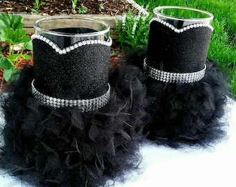 May include: Two glass candle holders decorated with black glitter fabric, silver rhinestone trim, and black tulle. The candle holders have a glamorous, formal appearance, suitable for weddings or special events.