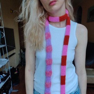 May include: A long, thin scarf with alternating stripes of pink and red. The scarf is knitted and has a textured look. The scarf is worn around the neck of a person.